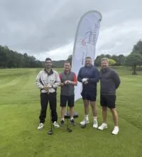 Image of four men on a golf course stood in front of a flag
